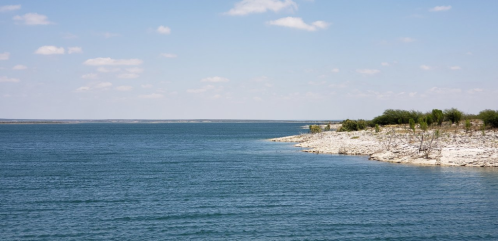 A serene lake with clear blue water, rocky shoreline, and a few trees under a bright sky with scattered clouds.