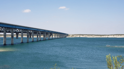 A long bridge spans over a calm blue lake, with clear skies and distant land visible in the background.