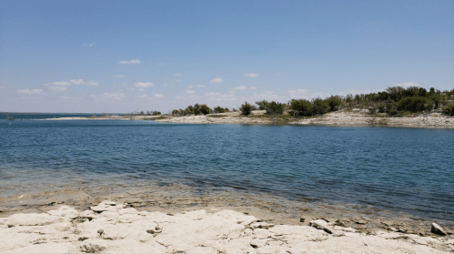 A serene lakeside view with clear blue water and rocky shores under a bright sky with scattered clouds.