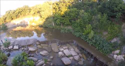 A serene river scene with a small waterfall, surrounded by lush greenery and rocky terrain under a golden sunset.