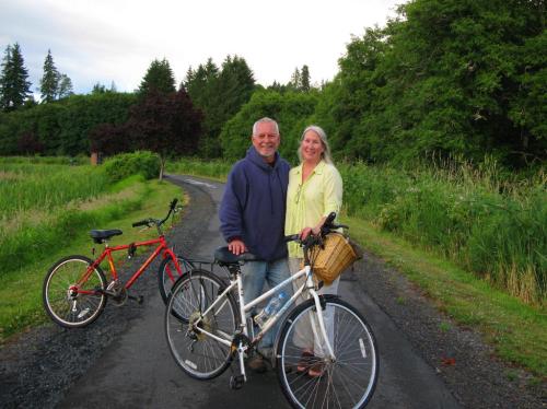 A smiling couple stands on a path with bicycles, surrounded by greenery and trees.