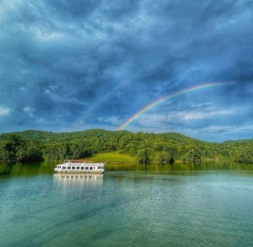 A serene lake scene with a boat and a vibrant rainbow arching over lush green hills under a cloudy sky.