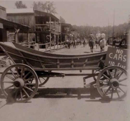 A vintage wagon on a dirt street with a "No Cars" sign, surrounded by wooden buildings and people on horseback.