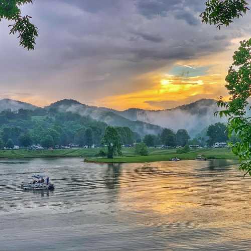 A serene landscape featuring a river, lush green hills, and a colorful sunset with mist rising in the background.