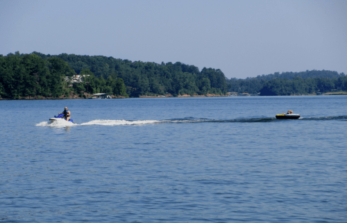 A person on a jet ski tows another person on an inflatable tube across a calm lake surrounded by trees.
