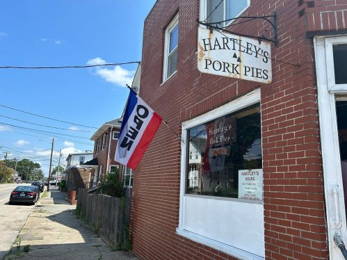 A brick building with a sign reading "Hartley's Pork Pies" and an open flag, set on a sunny street.