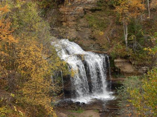 A serene waterfall cascades over rocks, surrounded by autumn foliage in vibrant shades of orange and yellow.
