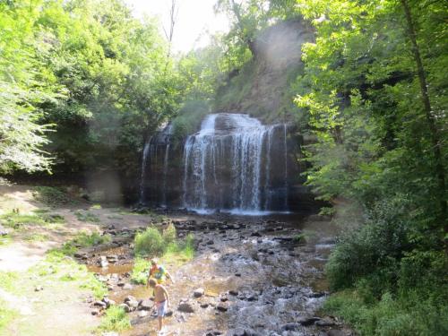 A serene waterfall cascades over rocks, surrounded by lush green trees and a person standing in the shallow water below.