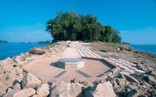 A scenic view of a rocky shoreline leading to a small island with trees under a clear blue sky.