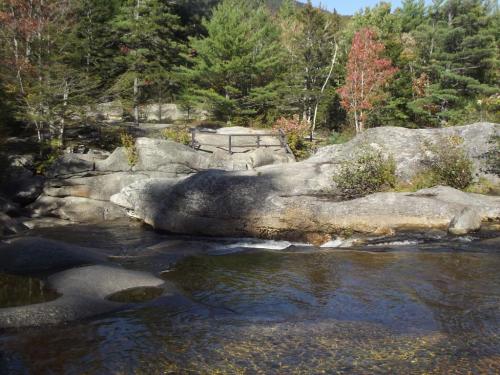 A serene river scene with smooth rocks, trees, and autumn foliage under a clear blue sky.