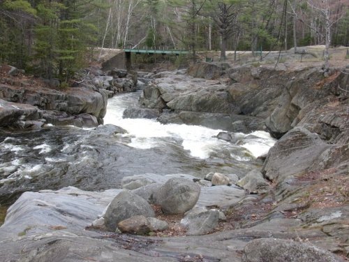 A rocky river landscape with flowing water and a bridge in the background, surrounded by trees.