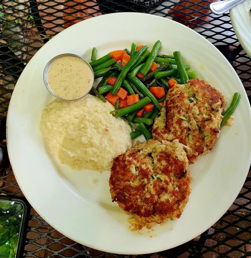 A plate with two crab cakes, creamy sauce, green beans, and mixed vegetables alongside a serving of grits.