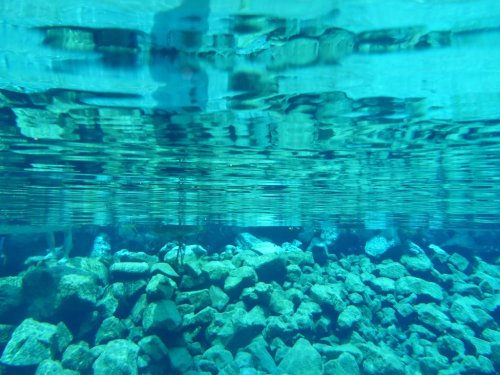 Underwater view of clear blue water with sunlight reflecting on the surface and rocky bottom visible below.