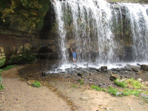 A person stands under a waterfall, surrounded by rocks and greenery, with water cascading down in front.