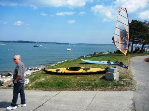 A man walks by a shoreline with kayaks and a windsurfing board, overlooking a calm blue sea and boats in the distance.