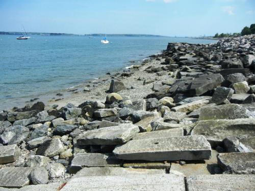 Rocky shoreline with scattered stones, calm water, and sailboats in the distance under a clear blue sky.