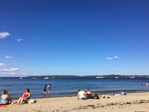 A sunny beach scene with people relaxing on the sand and boats in the calm water under a clear blue sky.
