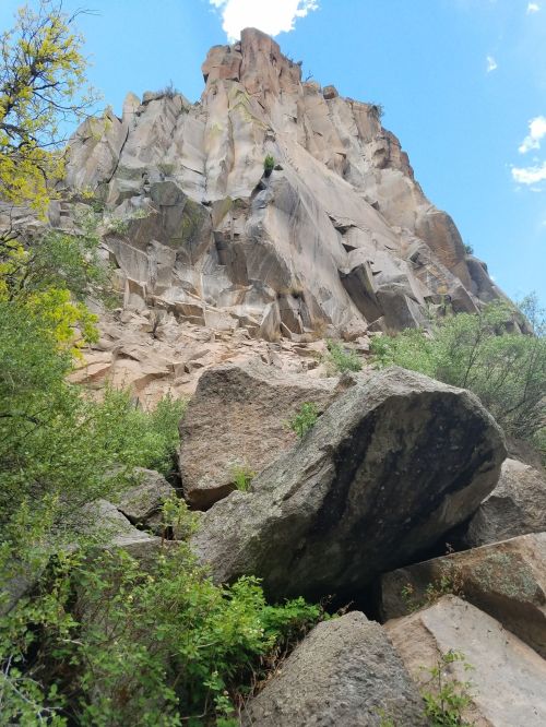 A rocky cliff rises against a blue sky, surrounded by greenery and large boulders at its base.