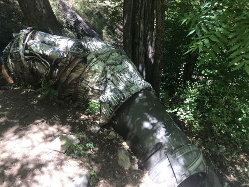 A large, curved metal pipe covered in camouflage fabric, lying on the ground among trees and greenery.