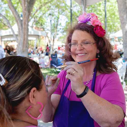 A smiling woman with a flower headband paints a child's face at an outdoor event. Trees and people are in the background.