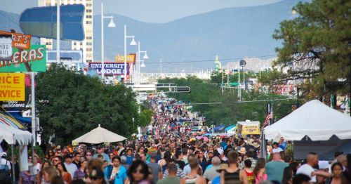 A bustling street filled with crowds of people, tents, and trees, set against a backdrop of mountains.