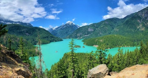 A panoramic view of a turquoise lake surrounded by lush green forests and mountains under a bright blue sky.