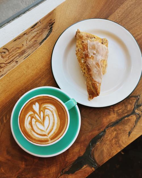 A latte with a heart design beside a glazed scone on a white plate, resting on a wooden table.