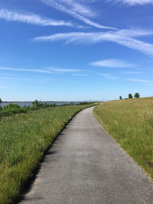 A winding path through green grass under a clear blue sky, leading towards a distant body of water.