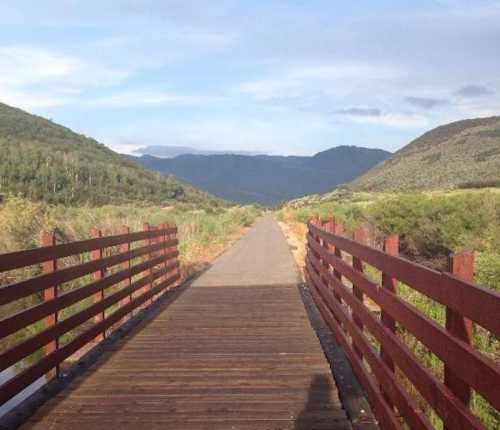 A wooden bridge leads through a scenic landscape of hills and greenery under a blue sky.