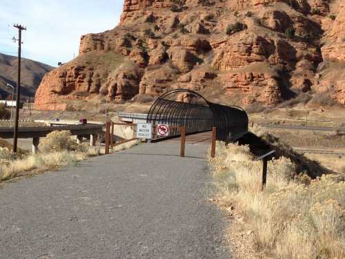 A pathway leads to a covered bridge over a highway, surrounded by rocky hills and dry vegetation. Signs indicate no motor vehicles.