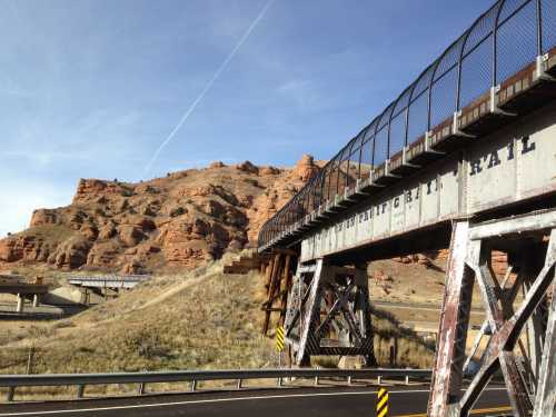 A weathered metal bridge spans a road, with rocky hills in the background under a clear blue sky.