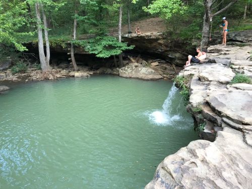 A serene natural pool surrounded by rocks and trees, with people relaxing nearby and a small waterfall flowing into the water.