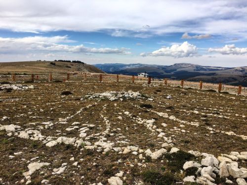 A rocky landscape with sparse vegetation, surrounded by mountains under a cloudy sky. A wooden fence outlines the area.