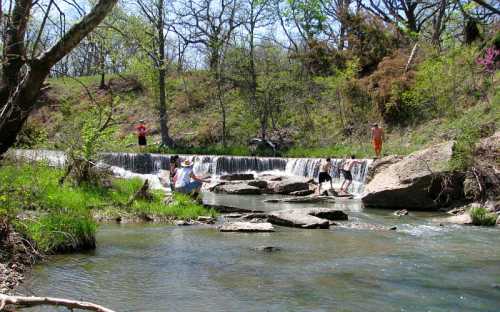 A serene landscape featuring a small waterfall, with people enjoying the water and surrounding greenery.