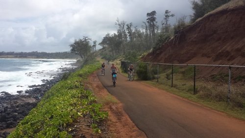 Bicyclists ride along a coastal path with lush greenery and ocean waves in the background.