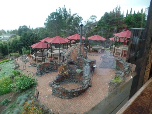 Outdoor dining area with stone features, red umbrellas, and lush greenery, viewed through a window.