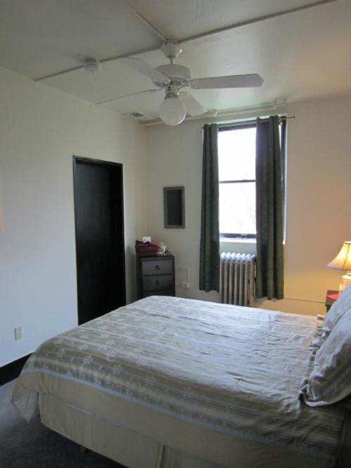 A simple bedroom with a bed, ceiling fan, window with green curtains, and a lamp beside the bed.