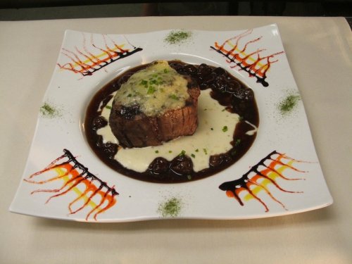 A gourmet dish featuring a beef filet topped with cheese, surrounded by a rich sauce and artistic plate decoration.