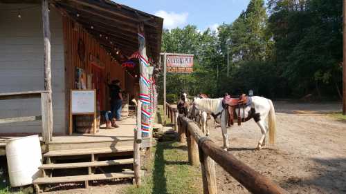 A rustic building with a porch, surrounded by trees, features horses tied up outside, ready for riding.