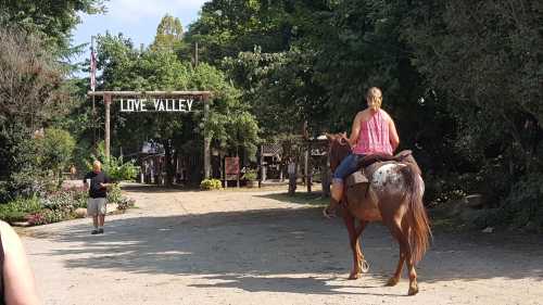 A person rides a horse towards a sign reading "Love Valley" in a rustic outdoor setting.