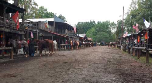 A rustic street lined with horses and wooden buildings, featuring flags and a muddy path under a cloudy sky.