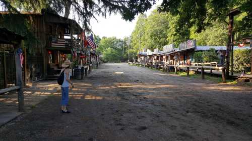 A person stands on a dirt path lined with rustic buildings and trees, under a clear blue sky.