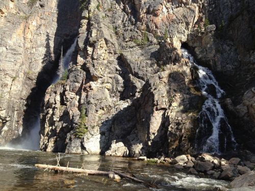 A scenic view of a rocky cliff with waterfalls cascading into a river below, surrounded by trees and sunlight.