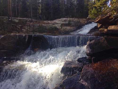 A serene waterfall cascades over rocks, surrounded by lush trees and a peaceful forest setting.