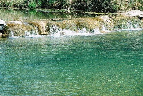 A serene water scene featuring a gentle waterfall cascading over rocks into a clear, calm pool. Lush greenery surrounds the area.