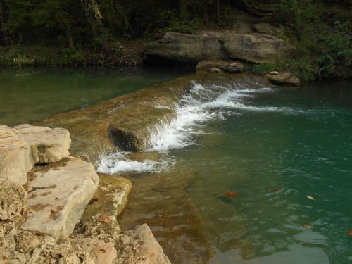A serene river scene with a rocky ledge and gentle water flowing over it, surrounded by lush greenery.