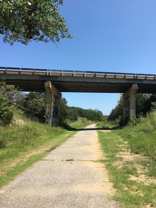 A clear path leads under a bridge, surrounded by greenery and blue sky.