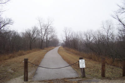 A foggy path lined with bare trees, leading into the distance, with a sign and a chain barrier at the entrance.