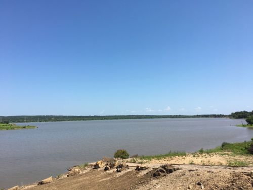 A calm lake under a clear blue sky, surrounded by greenery and a sandy shore in the foreground.
