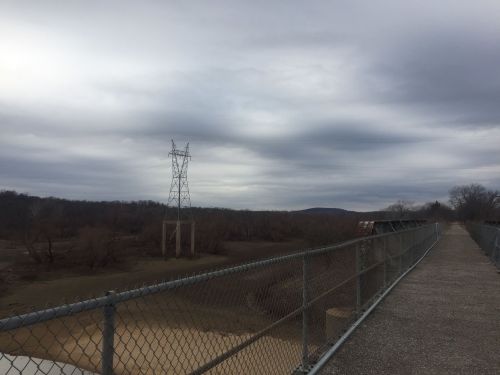 A gravel path with a chain-link fence, leading to a power tower against a cloudy sky and barren landscape.